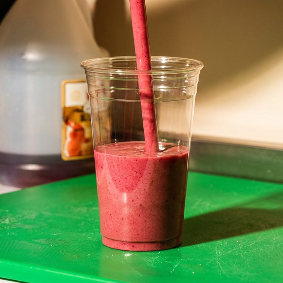 A vibrant pink smoothie is poured into a clear plastic cup on a green cutting board.