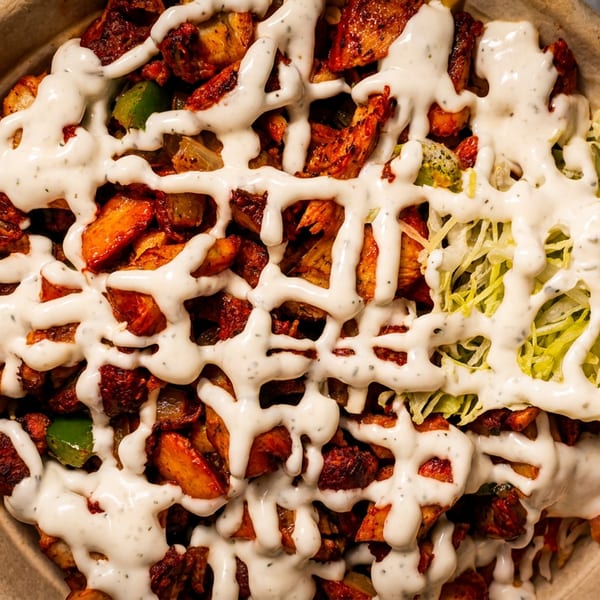 Close-up of a bowl filled with chicken, potatoes, green peppers, and shredded lettuce, drizzled with white sauce.